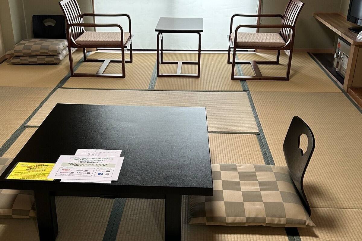 Une pièce avec un sol recouvert de tatamis, une table noire sur laquelle se trouvent des papiers, et deux chaises dotées de coussins à carreaux.