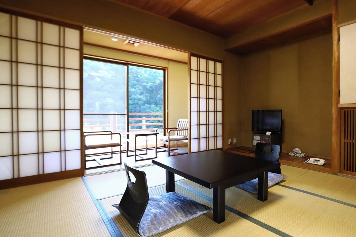 Une pièce de style japonais traditionnel, avec un sol recouvert de tatamis, une table basse, un téléviseur et une porte coulissante donnant sur un balcon.