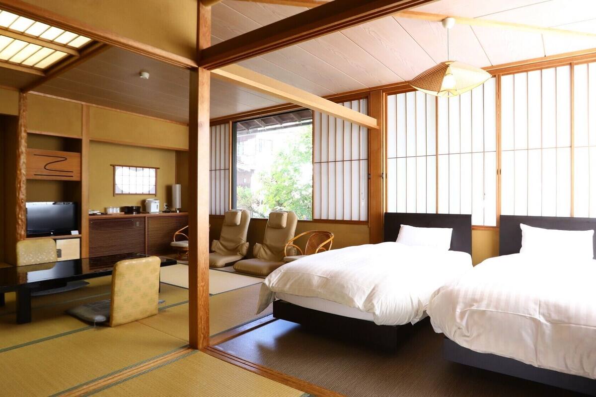 Une pièce de style japonais traditionnel, avec un sol recouvert de tatamis, une télévision, deux lits et une vue sur la verdure environnante.