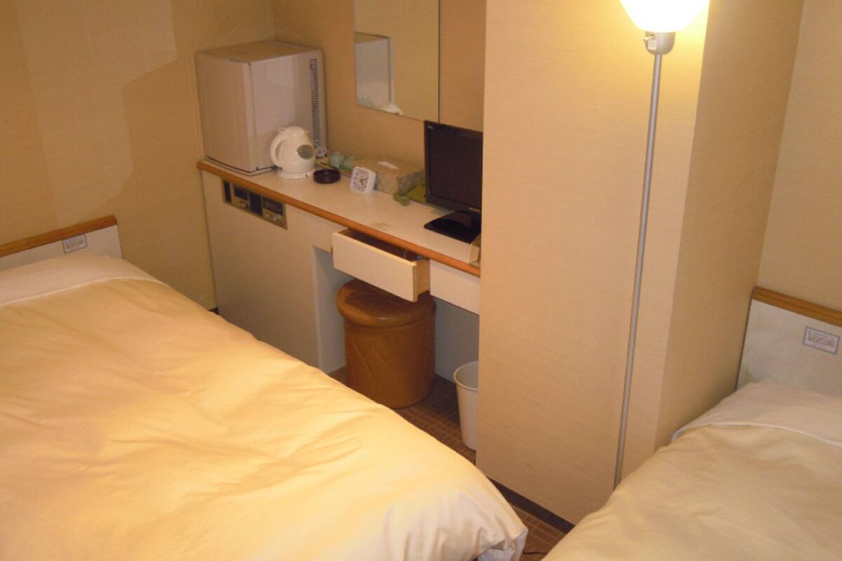 Ein kleines Hotelzimmer mit zwei Betten, einem Schreibtisch mit Computer, einer Lampe und einem Mülleimer.