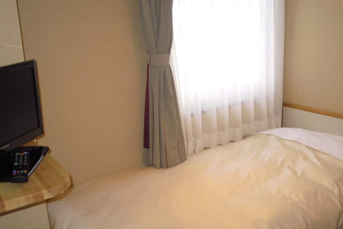 Ein Hotelzimmer mit Bett, Fernseher auf einem Holztisch und einem Fenster mit durchsichtigen Vorhängen.