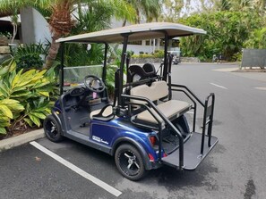 Un carrito de golf estacionado en un estacionamiento, con un edificio y follaje al fondo.