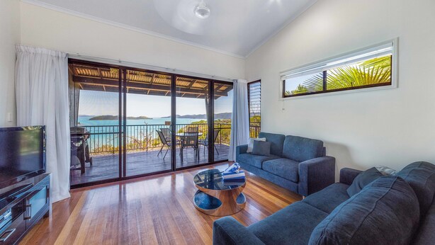 A spacious living room with a large sliding glass door leading to an outdoor deck, a comfortable sofa, a flat-screen TV, and a wooden coffee table. - Panorama 12 (Whitsundays)