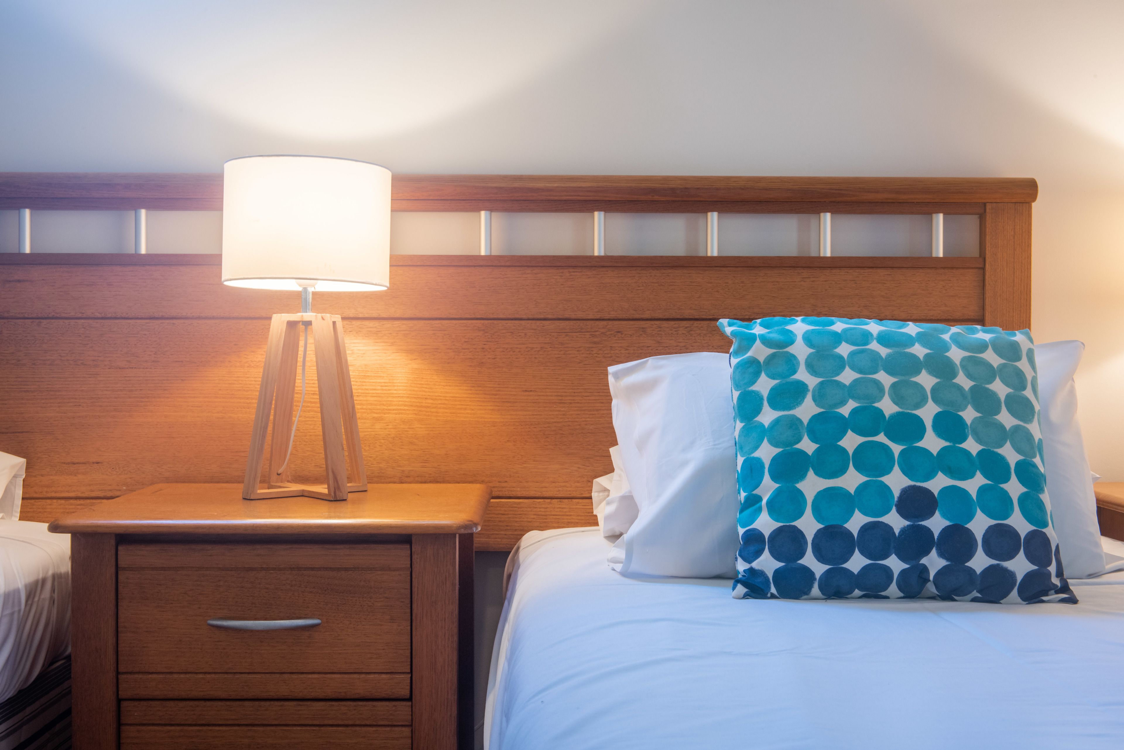 A neatly made bed with a white pillow and a blue polka dot pillow, a wooden nightstand with a lamp, and a wooden headboard.