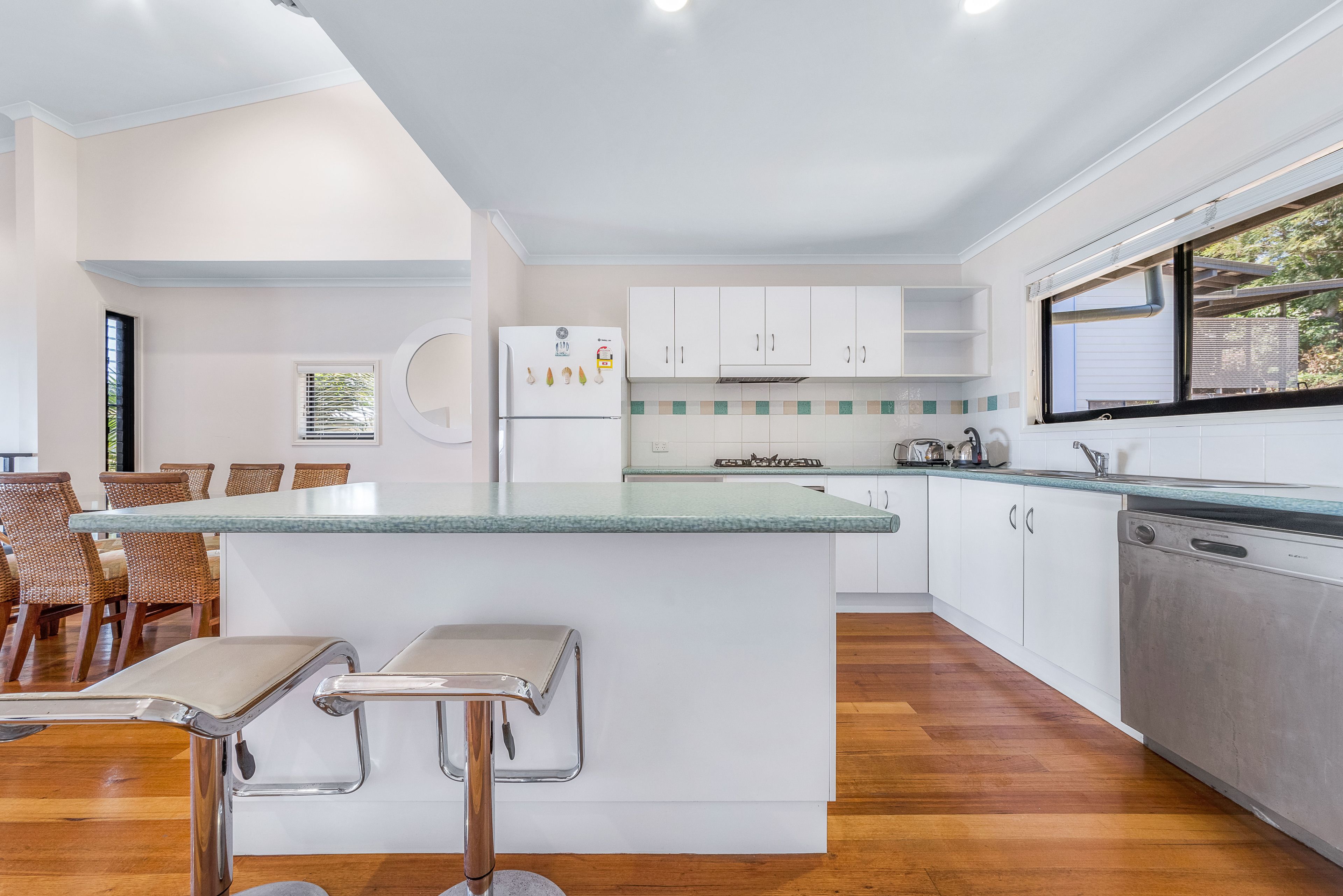 A modern kitchen with white cabinets, a central island with bar stools, a dishwasher, and a window with a view.