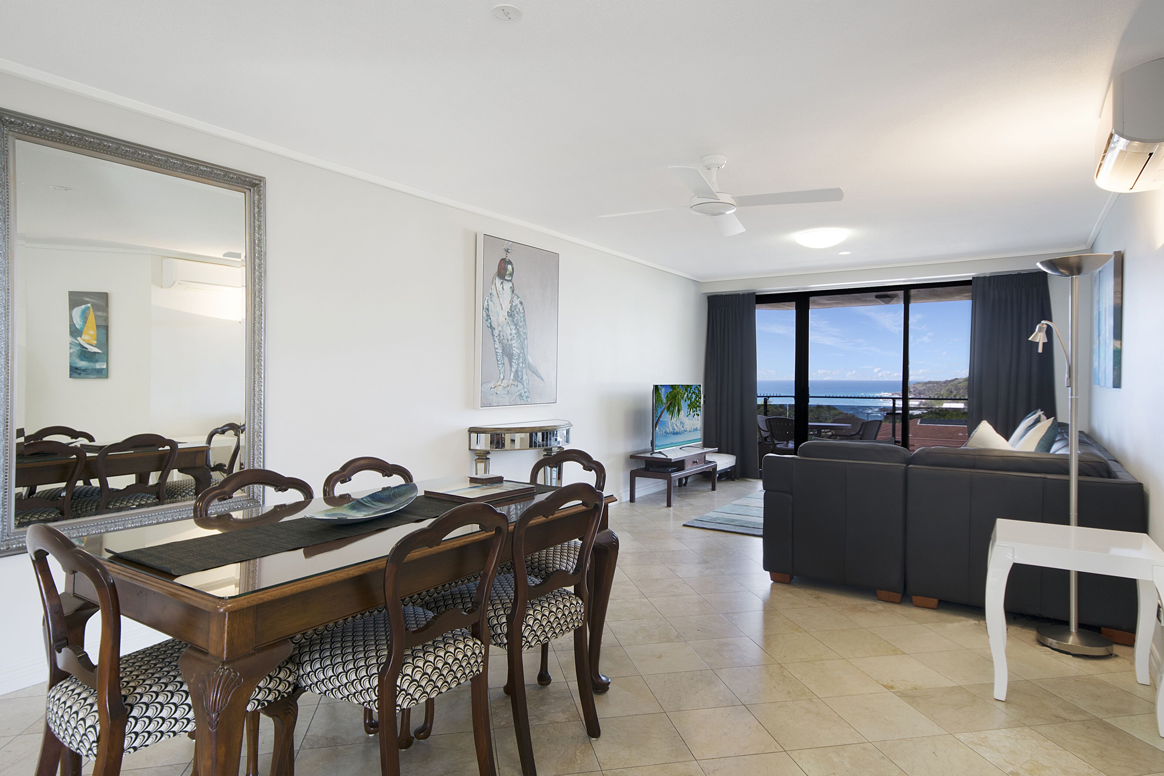 A modern living room with a dining area, a large window with a view, and a ceiling fan.