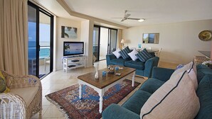Amplia sala de estar con televisor grande, asientos cómodos y vista al océano a través de un ventanal corredizo.