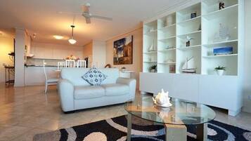 A modern living room with a white sofa, a glass coffee table, and a white shelving unit with decorative items.