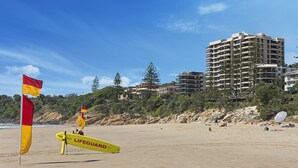Una playa con banderas de salvavidas, un quiosco de socorristas amarillo y un edificio de varios pisos al fondo.
