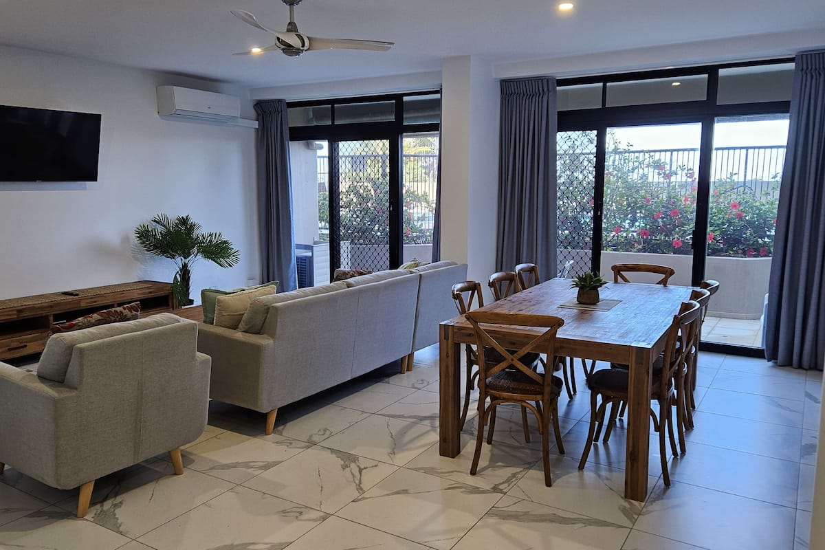Una sala de estar moderna con un sofá, una mesa de comedor de madera con sillas, un televisor montado en la pared y un ventilador de techo.