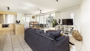 A modern living room with a grey sofa, a flat-screen TV, and a dining area with a wooden table and chairs. - The Cove Noosa (Noosa Heads)