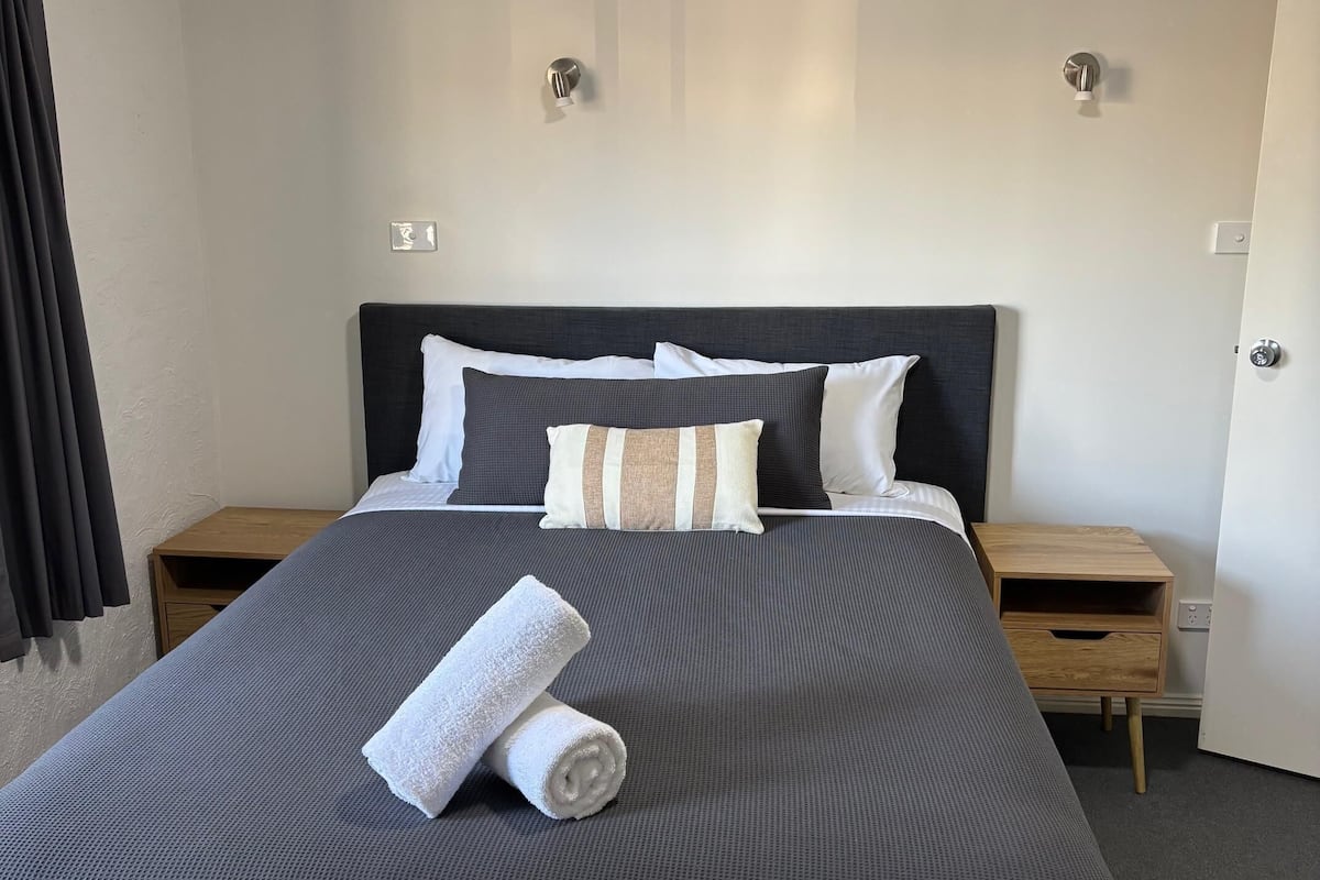 A neatly made bed with a dark headboard, white and striped pillows, and a rolled white towel on the bed. There are two wooden nightstands with drawers, a ceiling fan, and a white door.