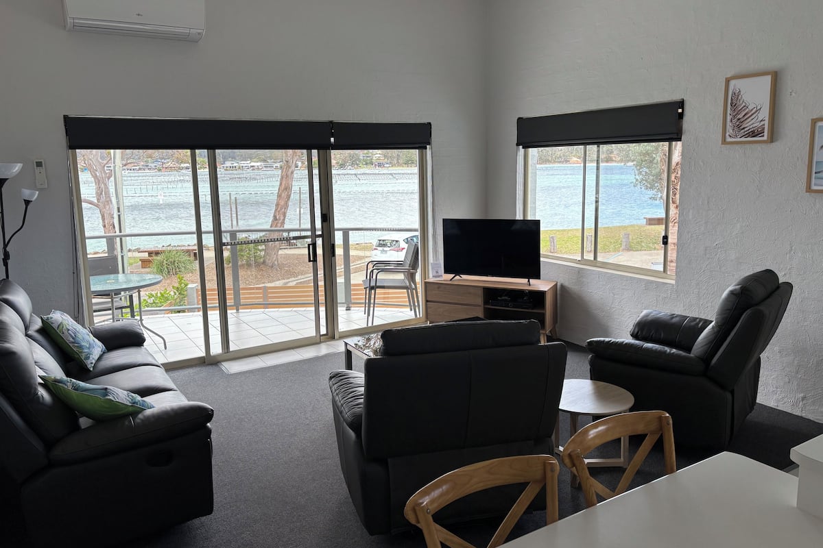 A living room with a TV, sofa, and chairs, large windows with a view of a body of water, and an air conditioning unit.