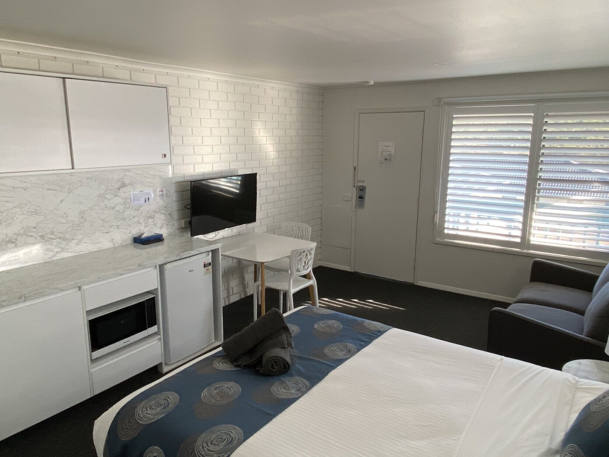 Habitación de hotel compacta con cama, una pequeña kitchenette, televisión, ventana con persianas y un sofá.