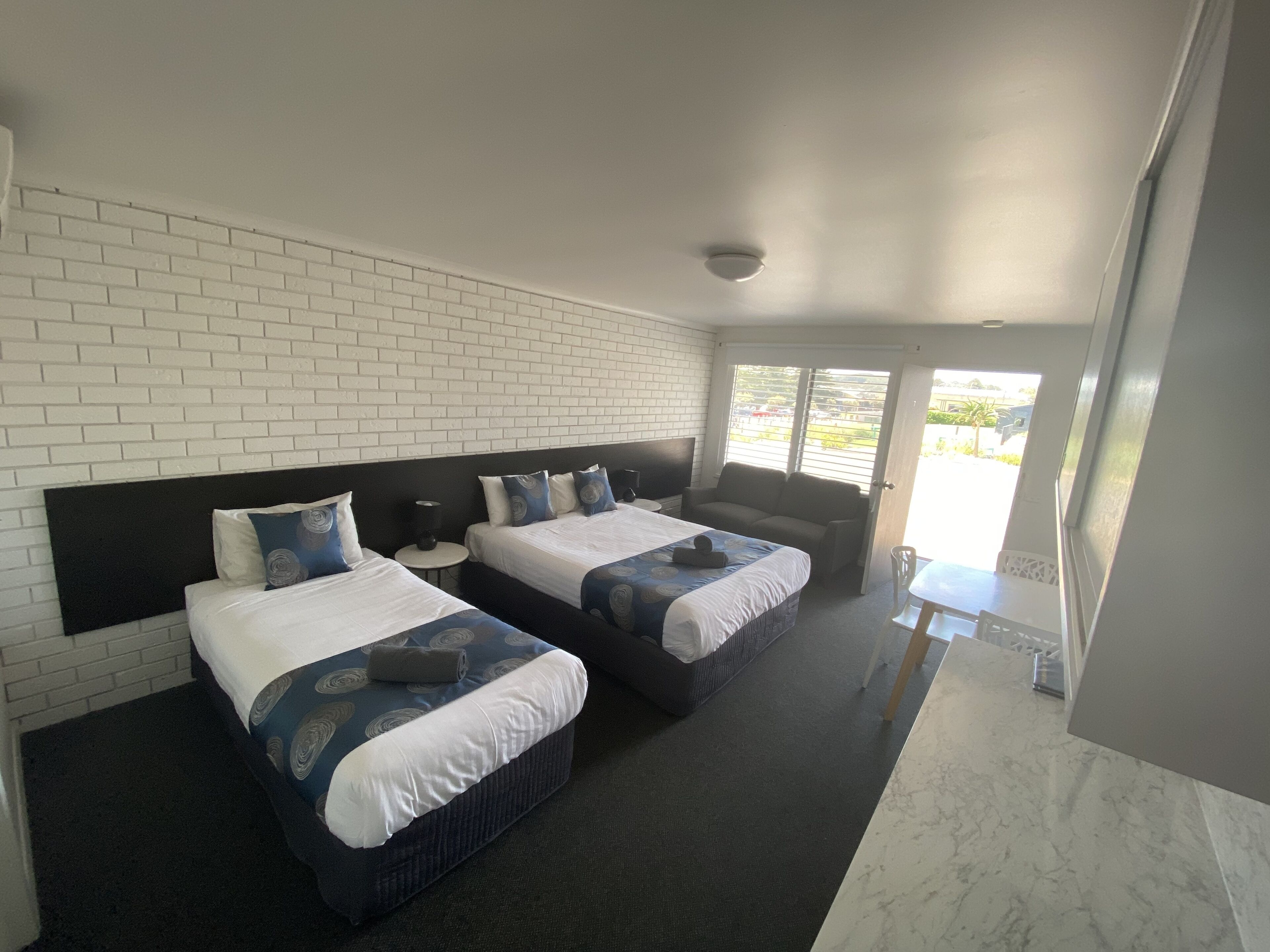 A hotel room with two beds, a white brick wall, a small table, and a view of the outdoors.