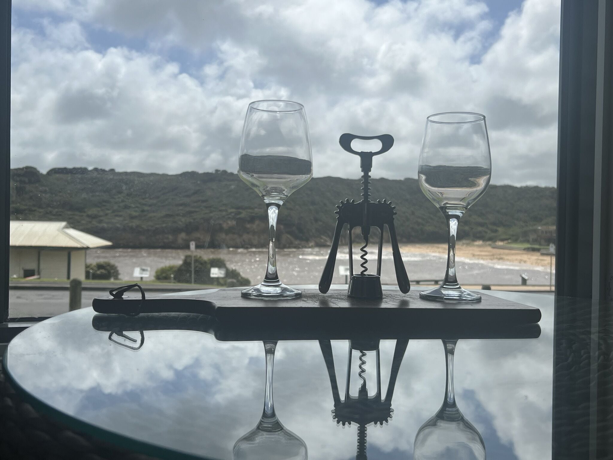 Ein Tisch mit zwei Weingläsern und einem Korkenzieher auf einer Glastischplatte, die einen Küstenblick mit Bergen und einem Gebäude im Hintergrund spiegelt.