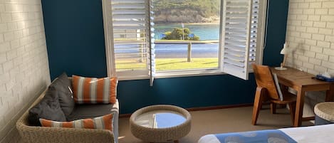 A bedroom with a bed, a wicker chair, a small round table, a sofa with cushions, and a window with white shutters.