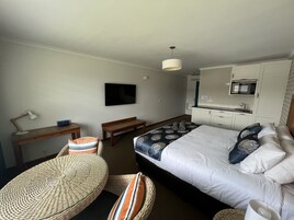 Una habitación de hotel con una cama, un escritorio con lámpara, televisor, kitchenette y una silla de mimbre.