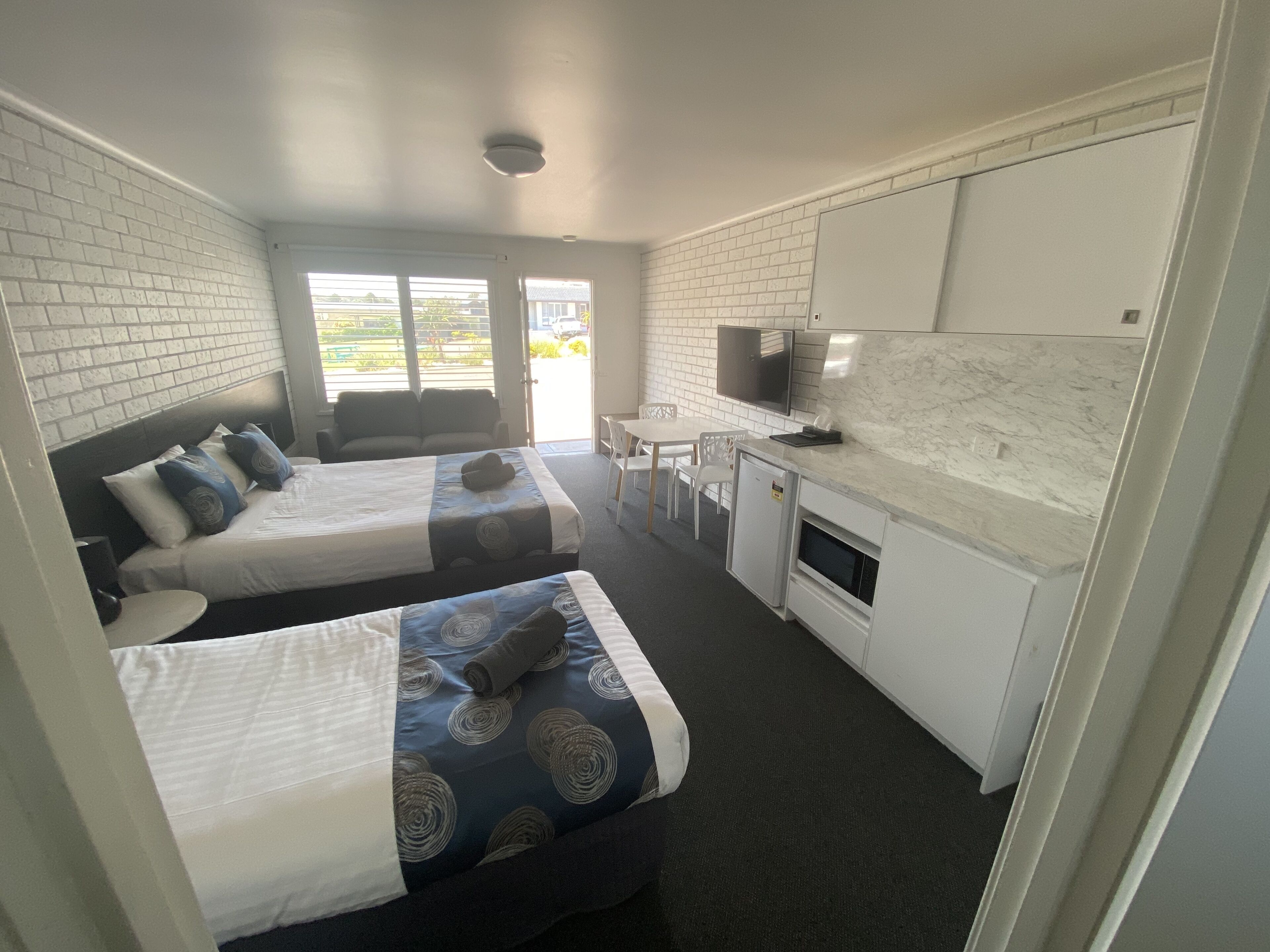 A hotel room with two beds, a kitchenette, a TV, and a view of the outside.