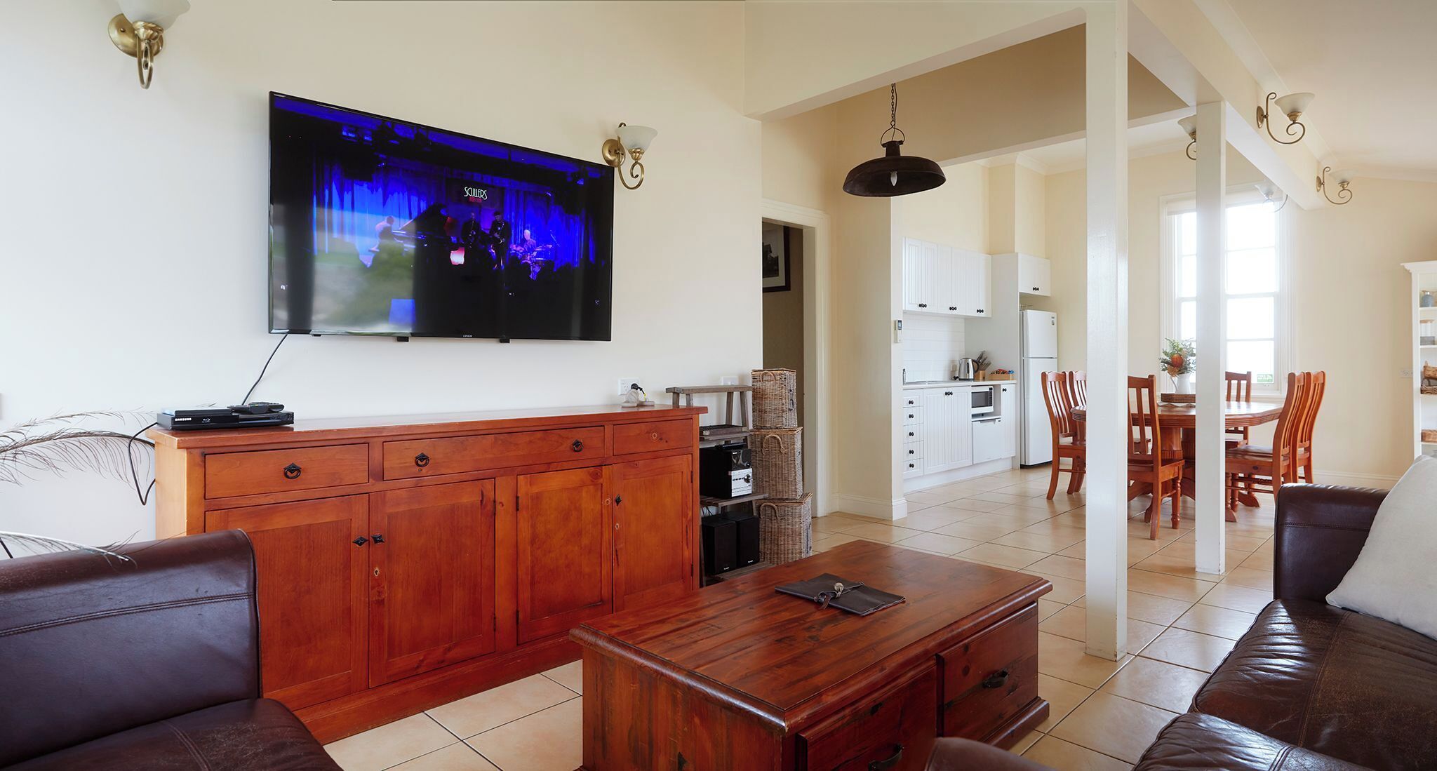 A living room with a wooden cabinet, a leather sofa, a TV mounted on the wall, and a kitchen area in the background.