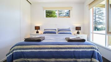 A bedroom with a large bed, two bedside tables with lamps, a window with a view of trees, and a small window above the bed.
