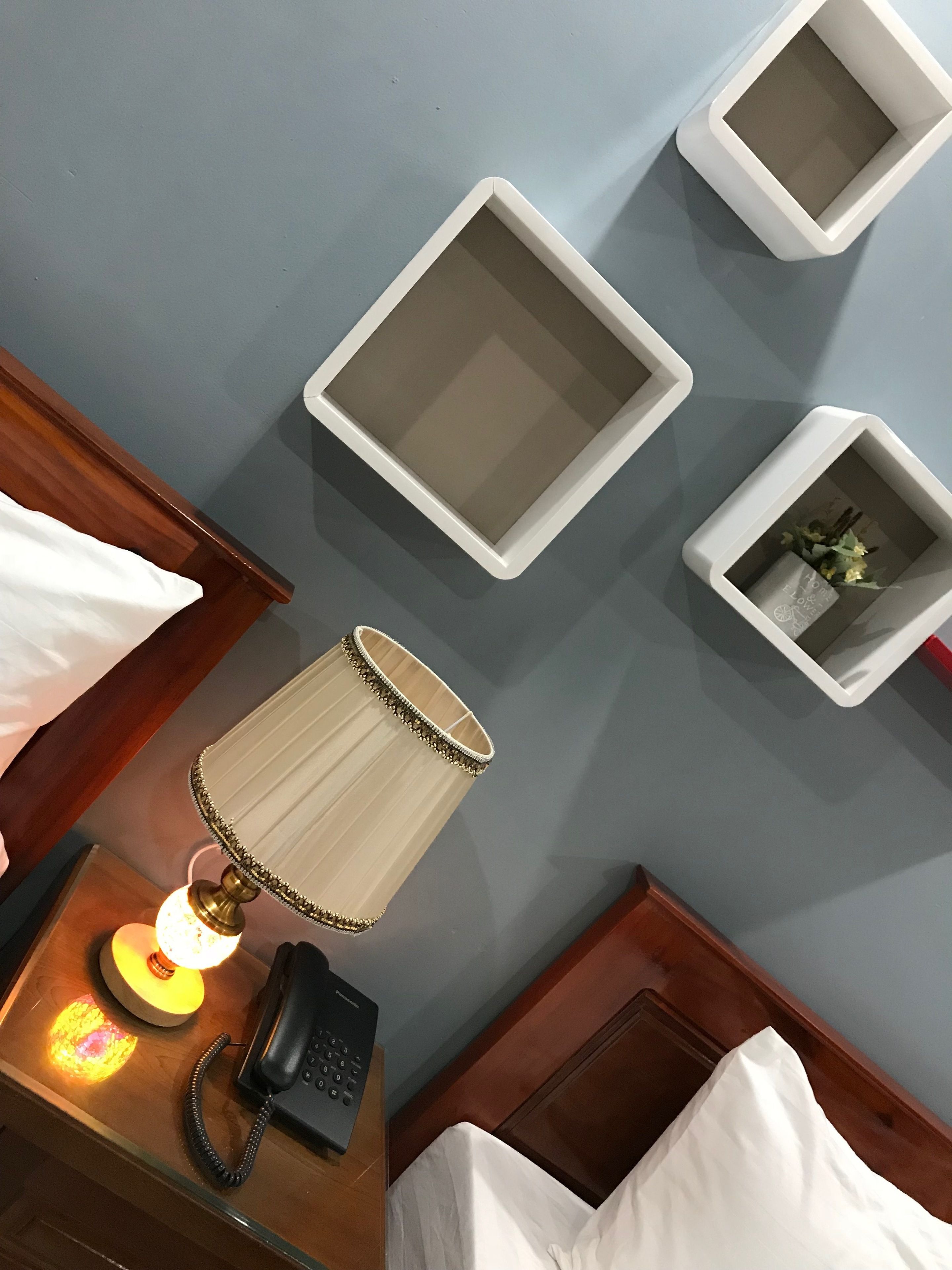 Habitación de hotel con una mesita de noche de madera, una lámpara, un teléfono y una cama con ropa blanca. Hay tres mesitas de noche blancas con adornos en la pared.