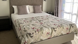 A bedroom with a large bed, a ceiling fan, a painting of birds, and a window with curtains. - Greenways Holiday Units (Tocumwal)