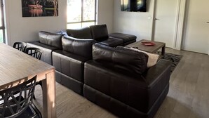A modern living room with a sectional sofa, a dining table with chairs, a ceiling fan, a flat-screen TV mounted on the wall, and a window providing natural light. - Greenways Holiday Units (Tocumwal)