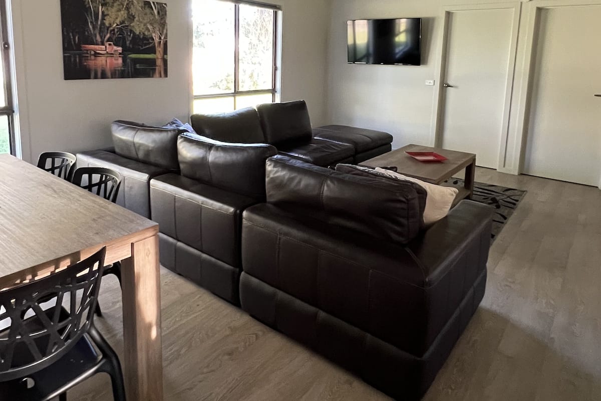 A modern living room with a sectional sofa, a dining table with chairs, a ceiling fan, a flat-screen TV mounted on the wall, and a window providing natural light.