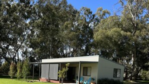 A small, single-story house with a flat roof, surrounded by a well-maintained lawn and tall trees. - Greenways Holiday Units (Tocumwal)