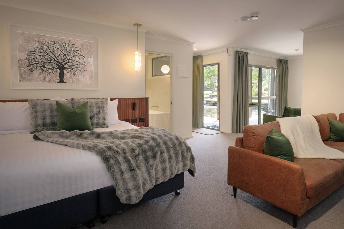 A modern hotel room with a bed, a sofa, a large window, and a painting on the wall.