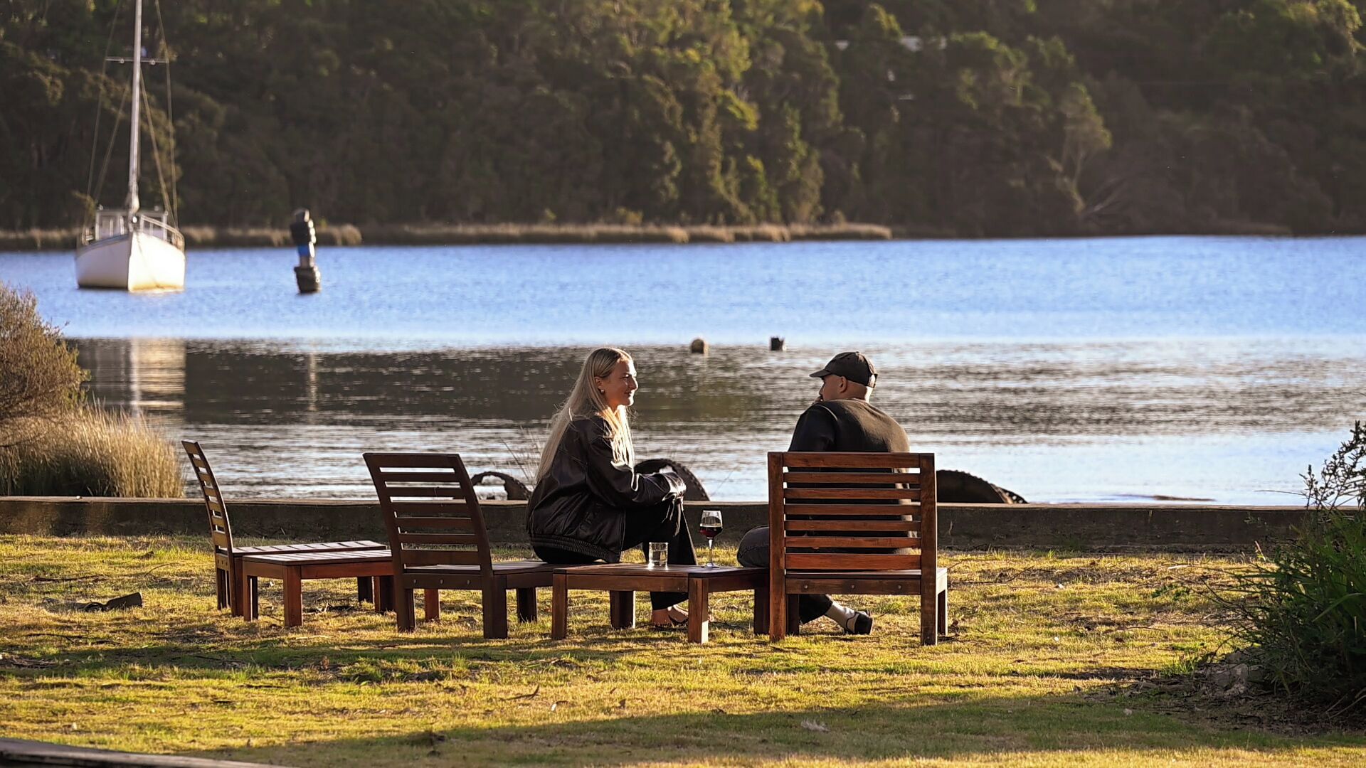 Duas pessoas sentadas em bancos de madeira à beira de um lago, com um veleiro ao fundo.