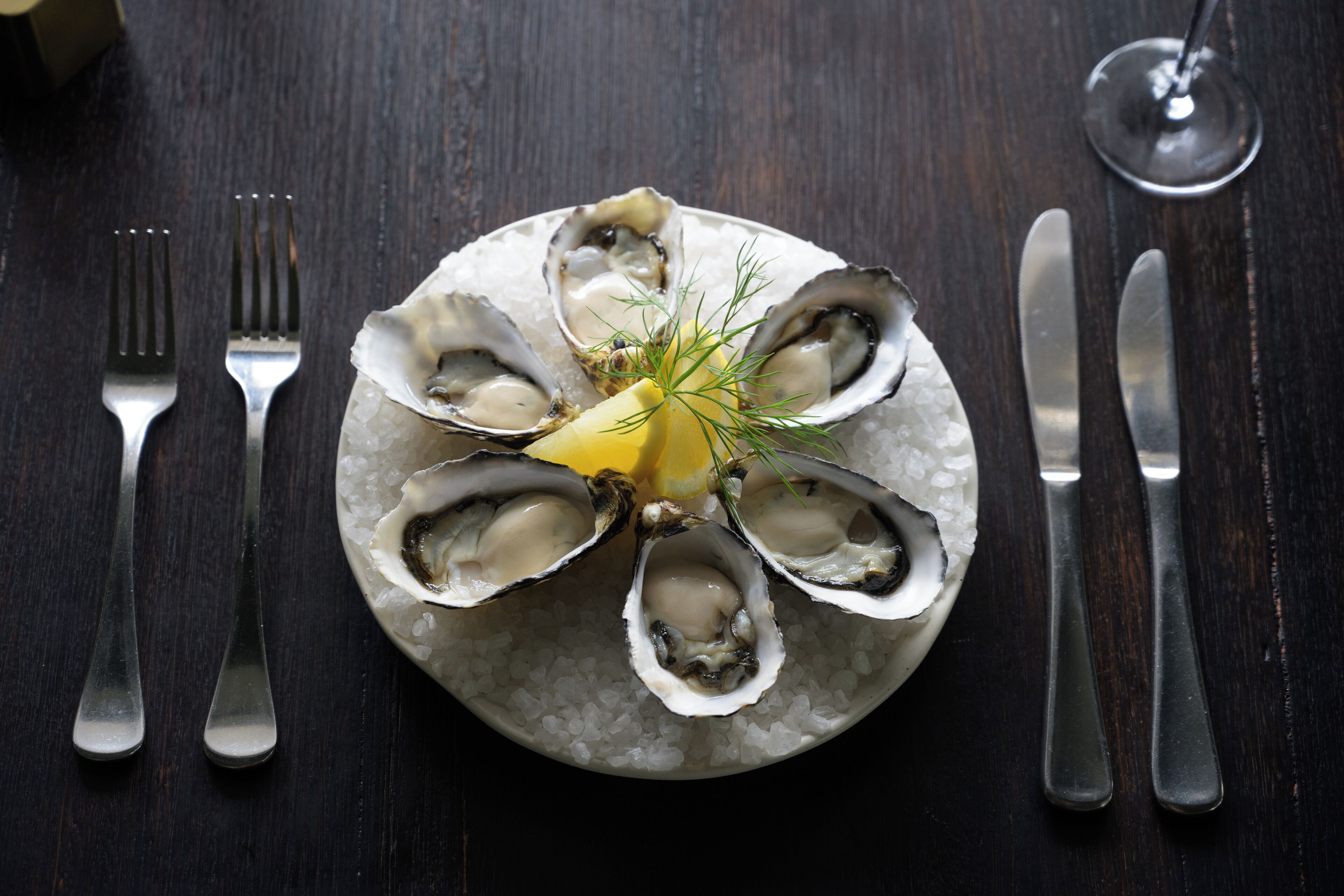 A plate of oysters on ice with a lemon wedge and dill garnish, accompanied by cutlery on a dark wooden table.