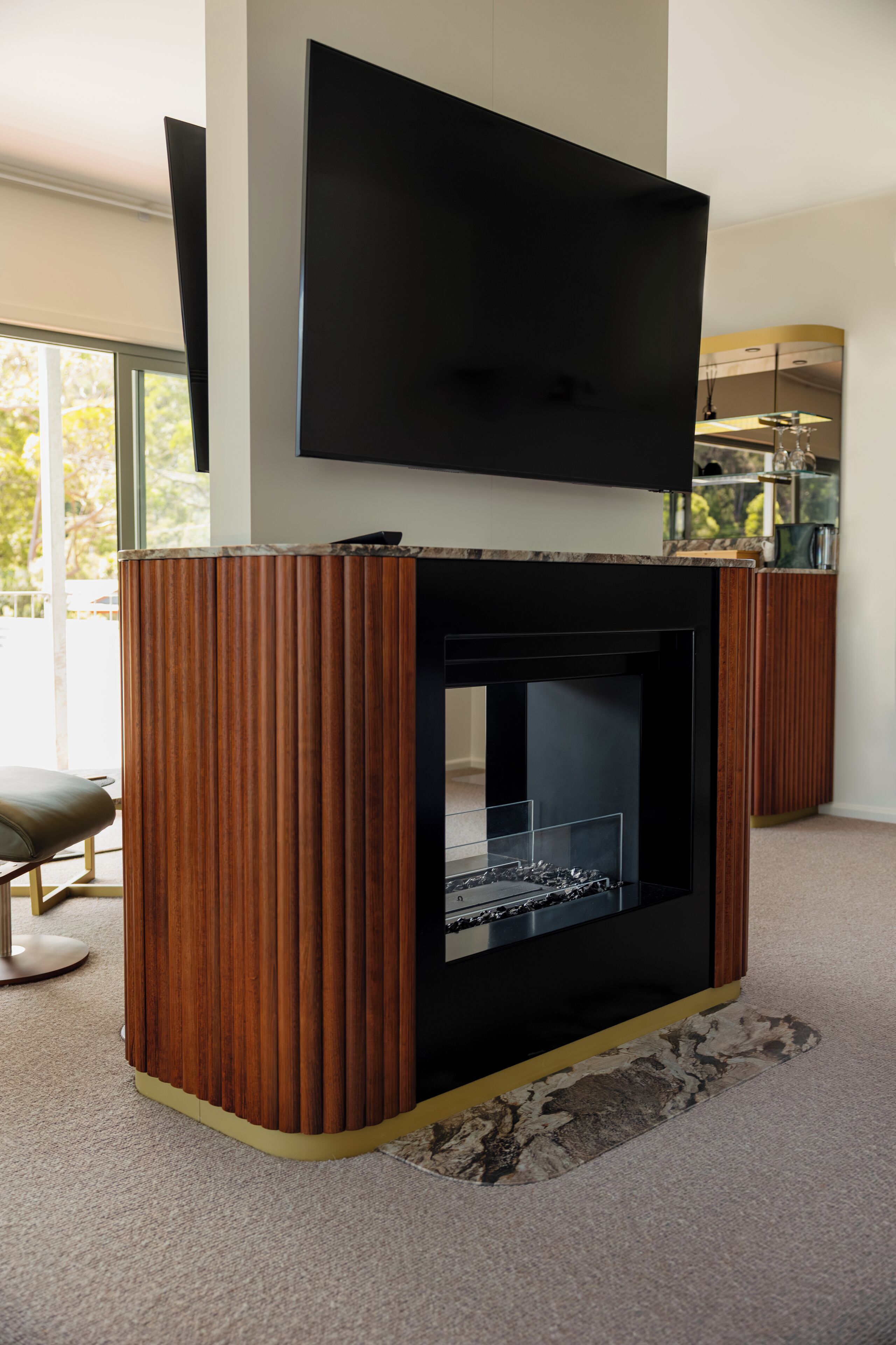 A modern living room with a fireplace, a flat-screen TV mounted above it, and a large window providing natural light.