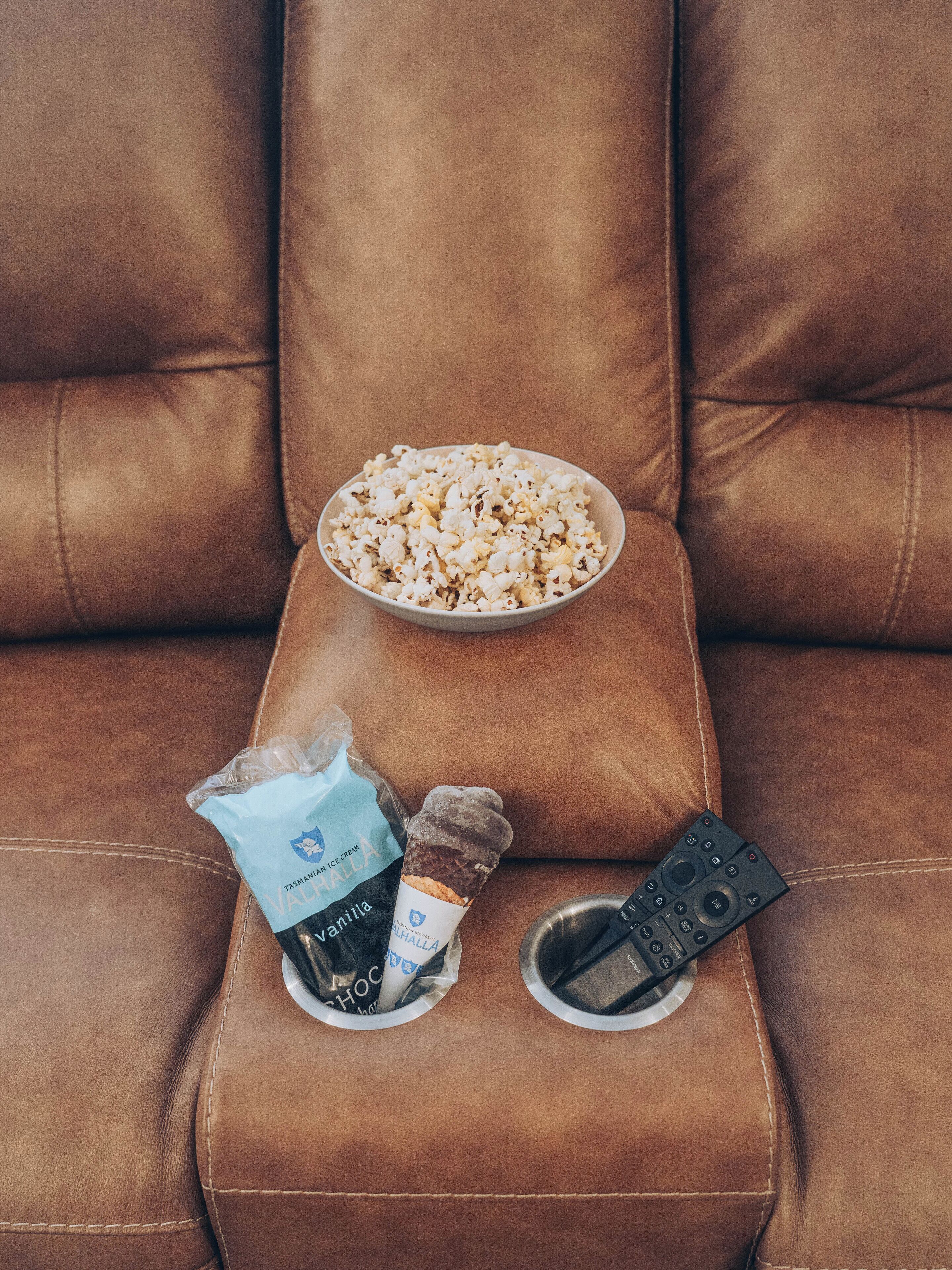 En brun lædersofa med en skål popcorn, en pose chokoladeis og en fjernbetjening.