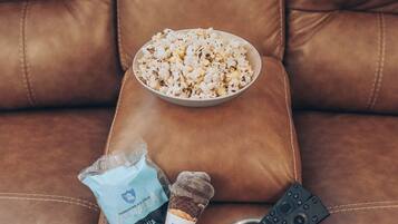 En brun lædersofa med en skål popcorn, en pose chokoladeis og en fjernbetjening.