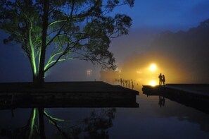A serene waterfront scene with a reflective water surface, a solitary tree with illuminated branches, and a couple standing on a dock facing the sunset.