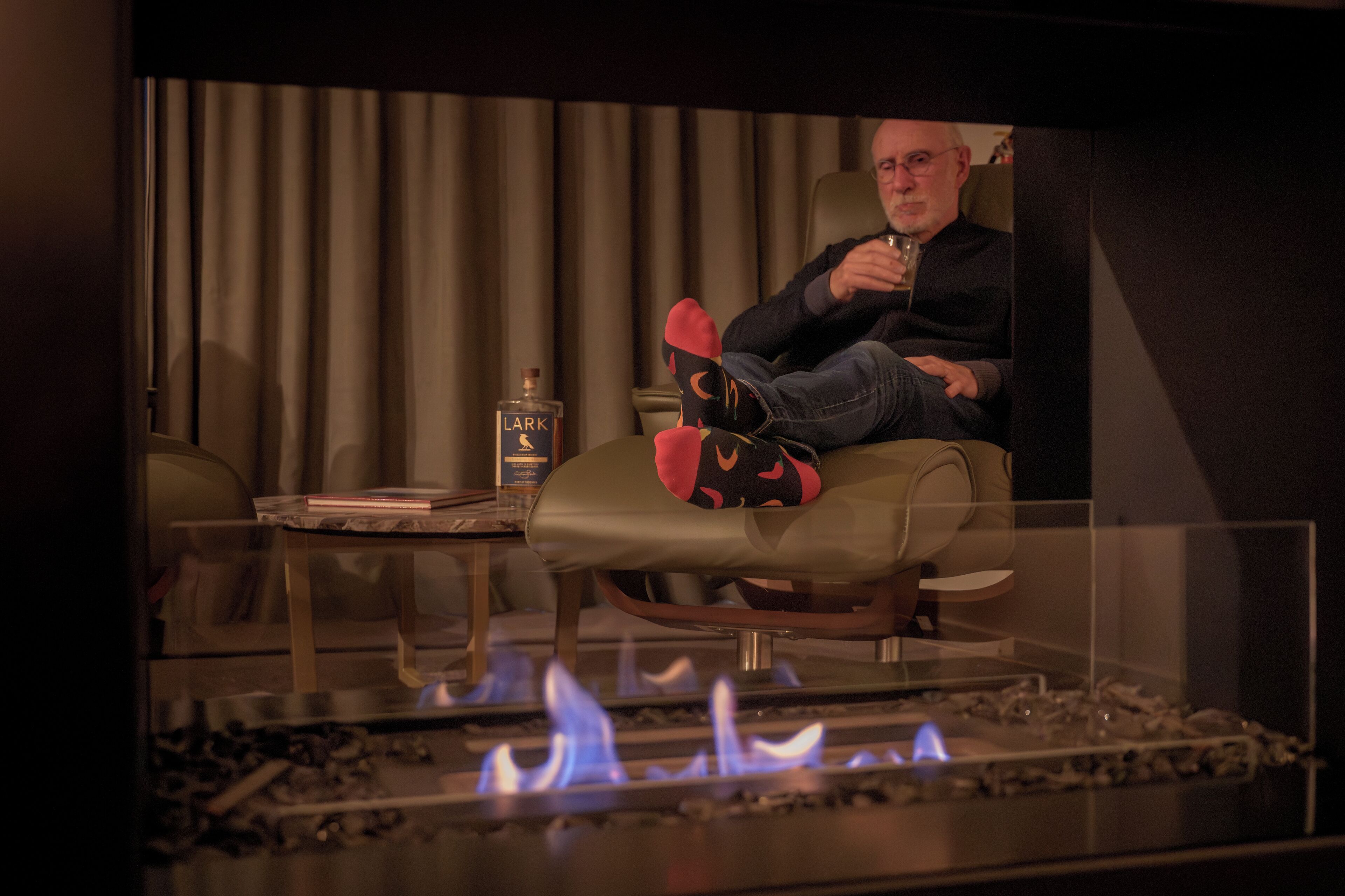A man seated in a leather armchair with a glass table in front of a fireplace.