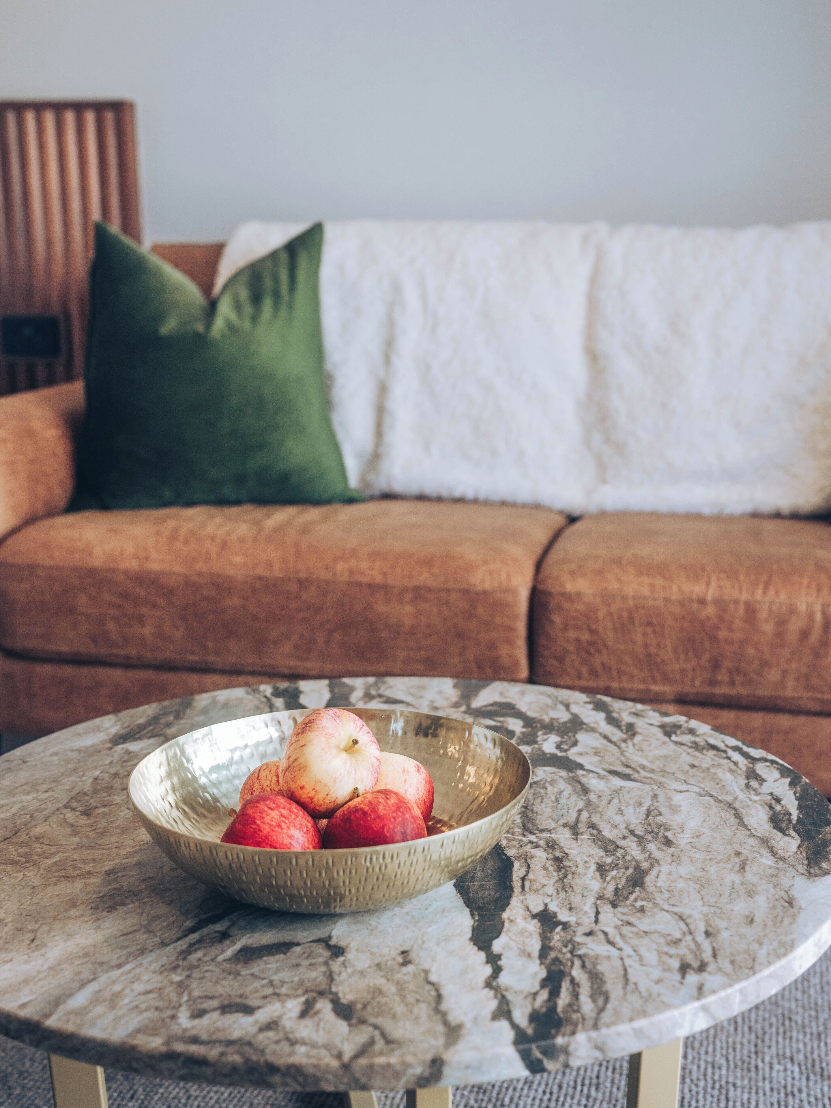A round table with a marble top, a bowl of apples, a green pillow, and a brown sofa in the background.