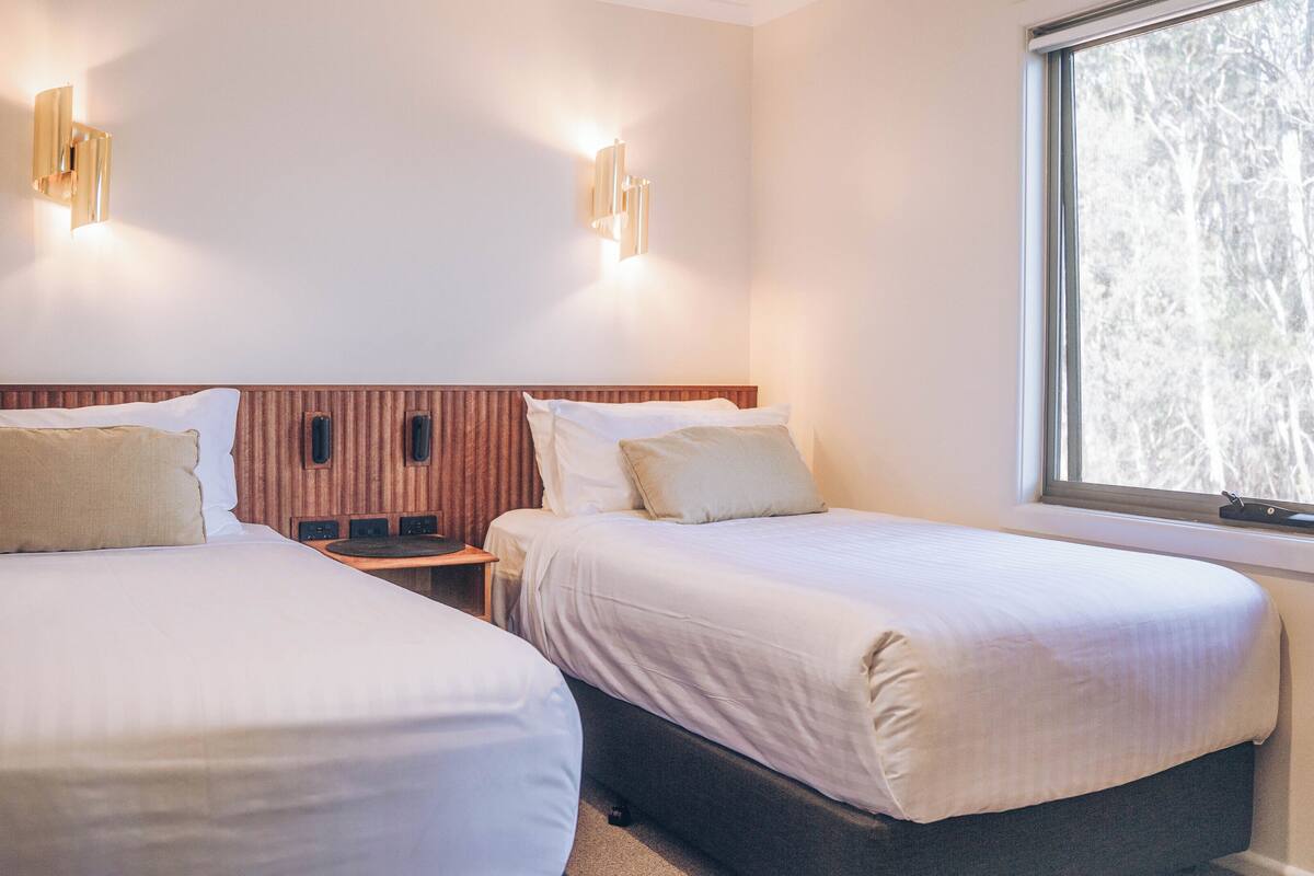 A hotel room with two single beds, a wooden headboard, bedside tables with lamps, and a window with a view of trees.