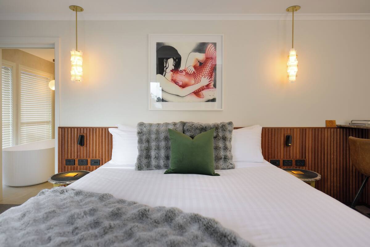 A hotel room with a large bed, two bedside tables, a green pillow, a framed artwork on the wall, and a wooden headboard.