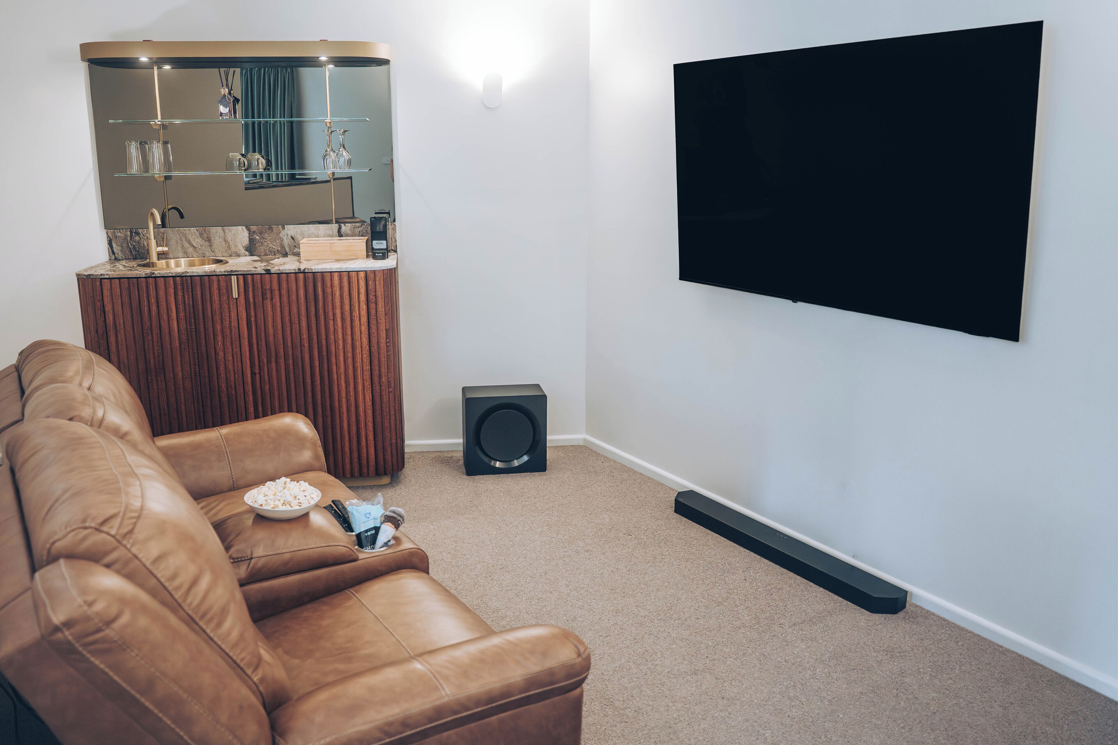A living room with a brown leather sofa, a TV mounted on the wall, a small black speaker, and a kitchen area with a marble countertop and wooden cabinets.