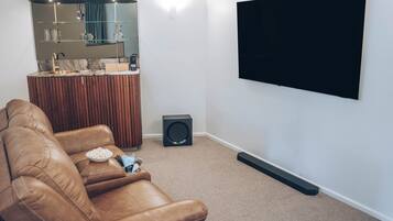 A living room with a brown leather sofa, a TV mounted on the wall, a small black speaker, and a kitchen area with a marble countertop and wooden cabinets.