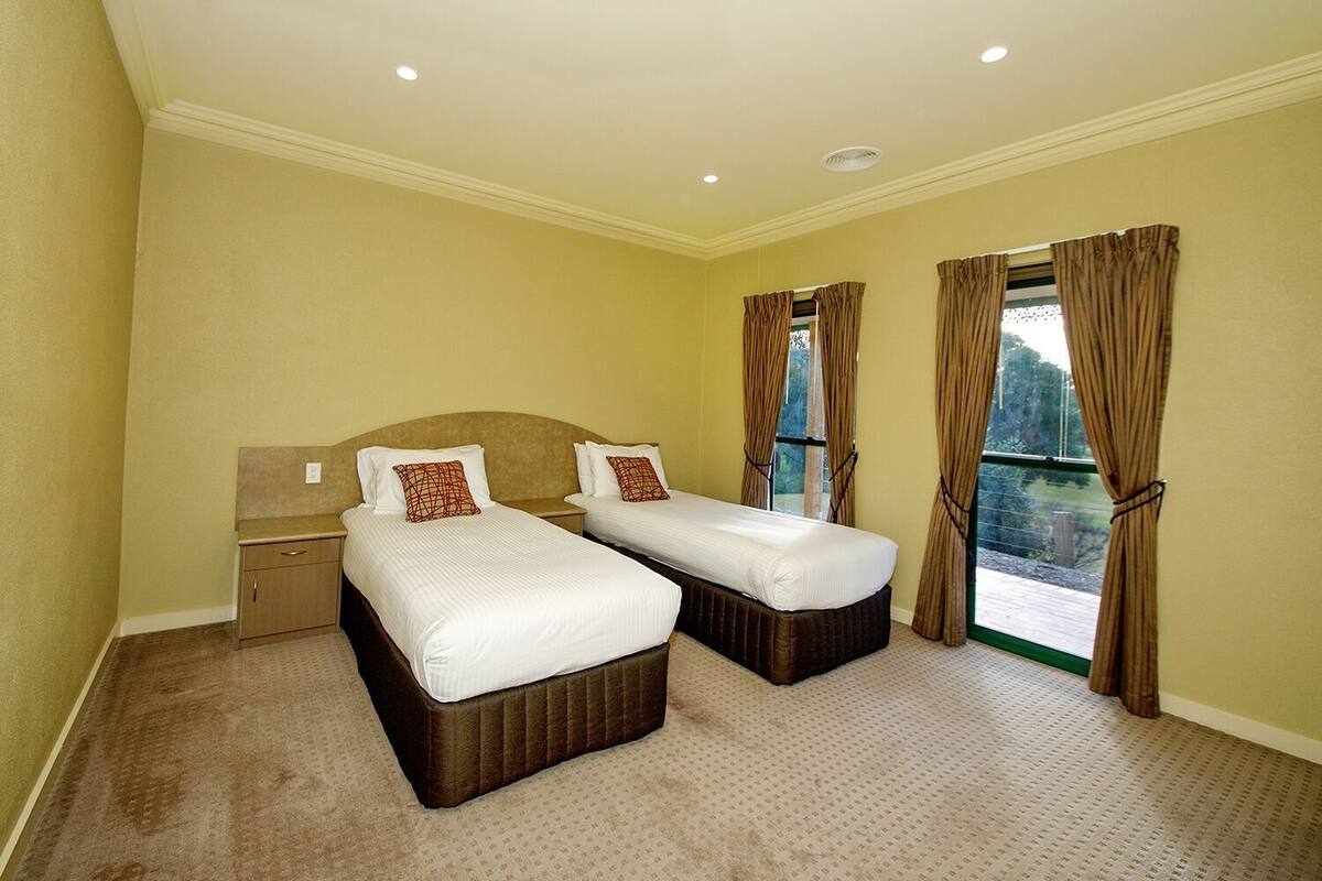A hotel room with two beds, a nightstand, a window with curtains, and a view of the outdoors.