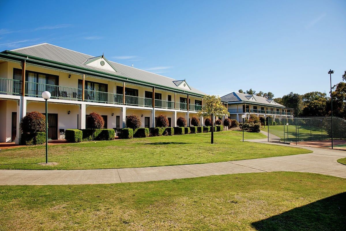 A hotel with a row of rooms, a green lawn, and a fenced sports area.