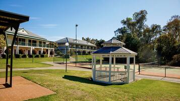 A gazebo in a well-maintained courtyard with a tennis court, surrounded by a fence and a building with a green roof.