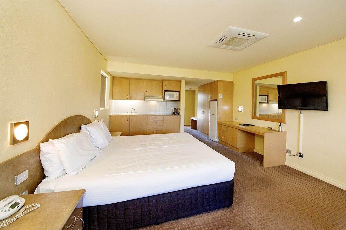A hotel room with a large bed, a desk with a telephone, a kitchenette, and a television mounted on the wall.