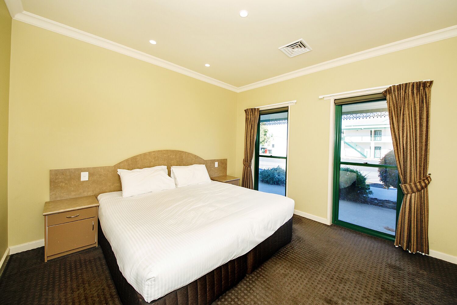 A hotel room with a bed, a nightstand, a window with curtains, and a view of the outside.
