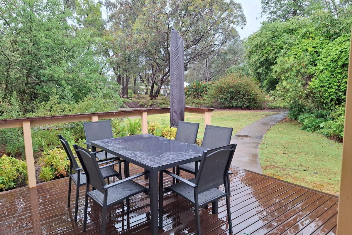 A covered outdoor deck with a table and chairs, surrounded by lush greenery and trees.