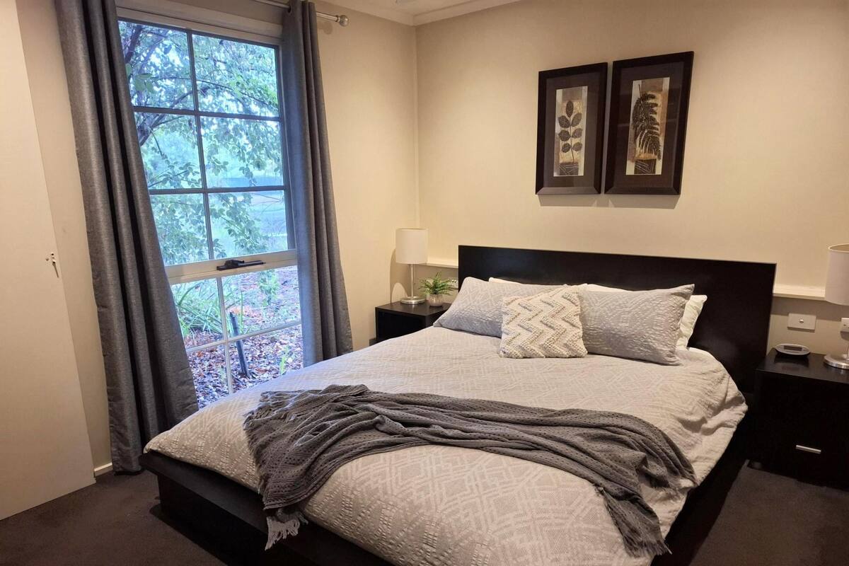 A neatly made bed with a gray blanket and pillows, a bedside table with a lamp, and a window with curtains.